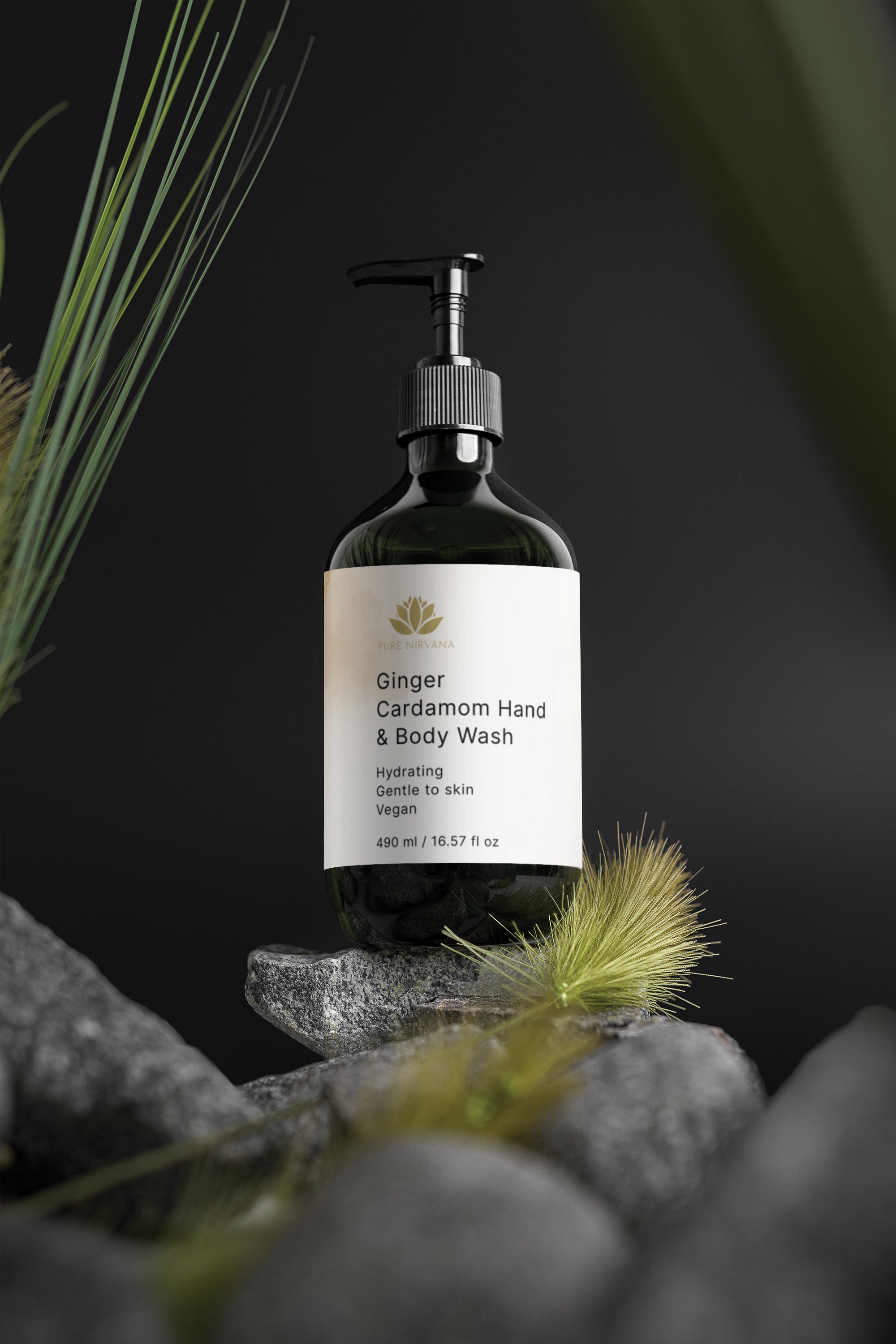 A dark amber pump bottle of Ginger Cardamom Hand & Body Wash stands on stacked gray stones with blurred green grass in the foreground and background against a dark backdrop.